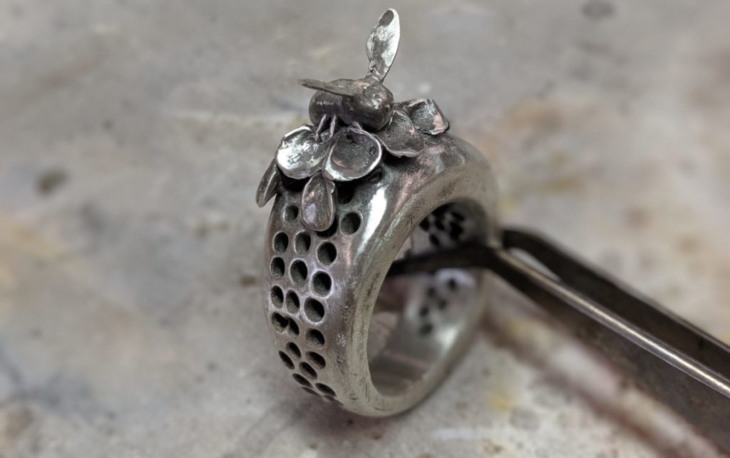 image of a silver ring held up with a pair of tweezers. On the ring there is a metal flower and a bee